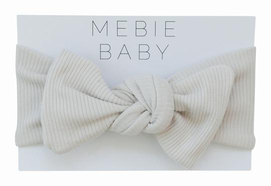 White background with Vanilla Organic Cotton Ribbed Head Wrap by Mebie Baby, in its packaging. Head wrap is cream ribbed with a knot bow.