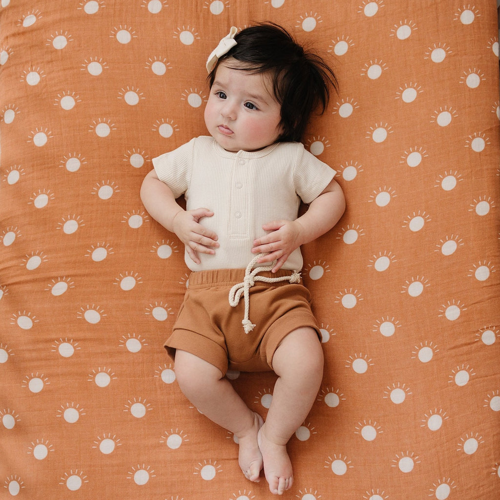 Vanilla Organic Bodysuit by Mebie Baby, paired with rust Mebie Baby Shorts and a Vanilla hair bow, with a sun and rust coloured blanket. A baby is wearing the outfit. 