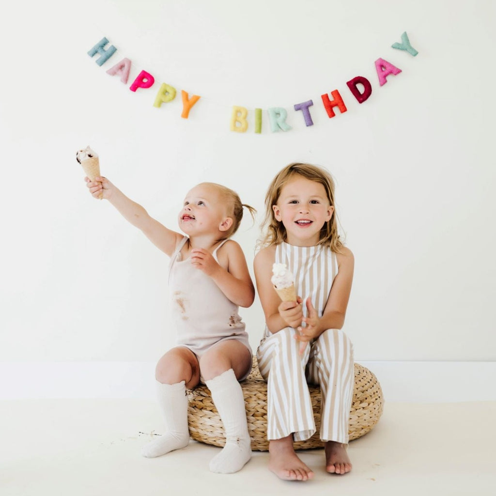 White wall with 2 little girls sitting on a rattan pout, with the Felt Happy birthday Garland by The Whimsical Woolies hanging on the wall.