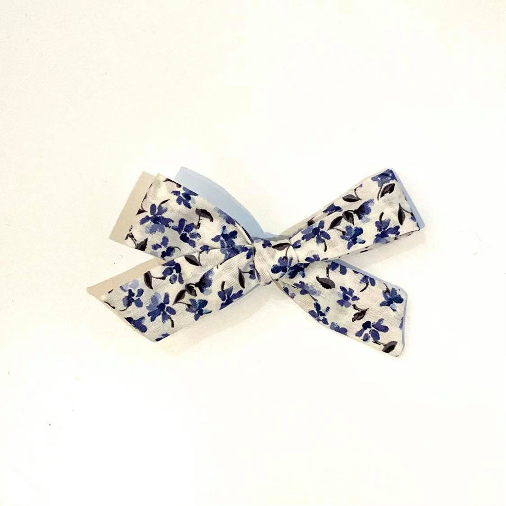 White background with the Blue Floral School Girl Bow by Wild and Free Design Co. Bow is a thin bow in white, with a blue floral pattern.