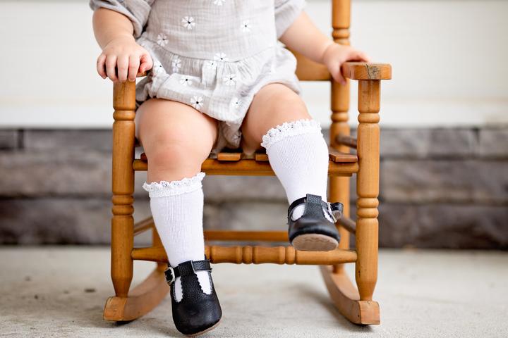 Baby sitting on a little wood rocking chair, wearing a romper, and a pair of Lace Knee High Socks in White by Little Stocking Co.