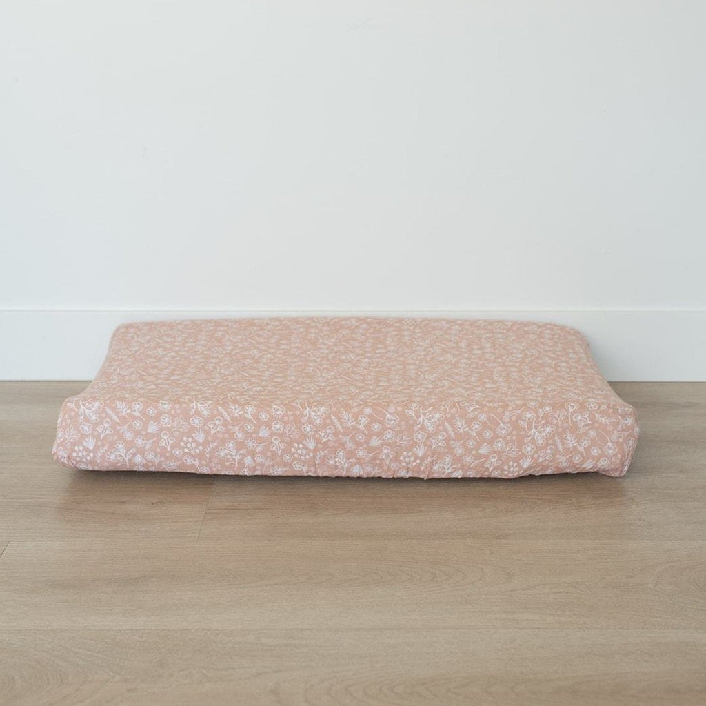 White wall with a light wood floor, and a change pad with a Wildflower Changing Pad Cover by Mebie Baby. Cover is pale pink with white wildflowers all over.
