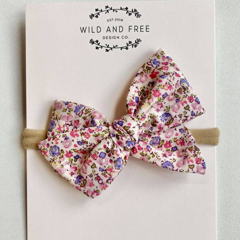 White background with the Blush Floral Pinwheel Bow by Wild and Free Design Co. on its packaging. Bow is a white colour with pink and purple floral pattern.