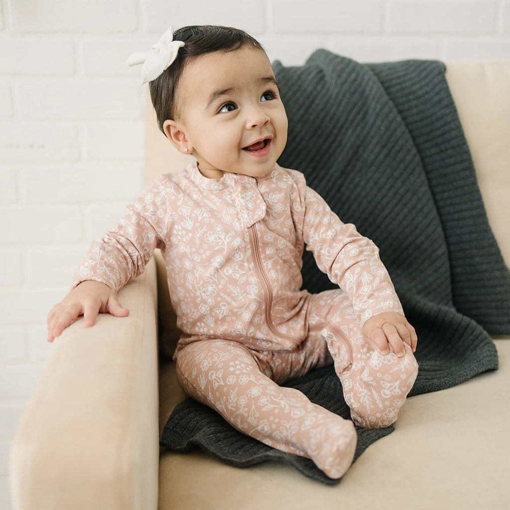 Baby girl sitting on a cream leather couch, wearing the Wildflower Zipper One-Piece by Mebie Baby.