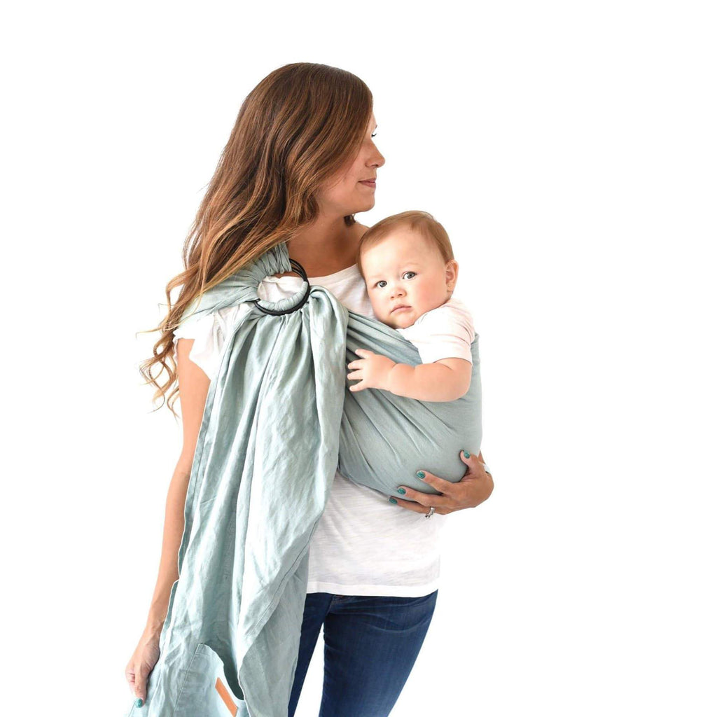 White background with woman standing, looking to the side, wearing her baby in a Ring Sling in Willow by Kyte Baby. Ring Sling is a blue colour.