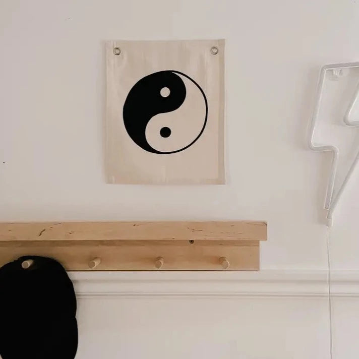 Yin Yang Wall Banner by Imani Collective, hanging on a white wall. Below it is a wooden shelf hook with a black hat hanging off one of the hooks. To the right, there is a neon bolt sign. 