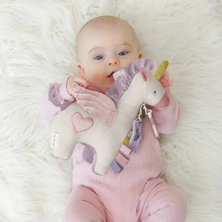 Baby model holding Bitzy Bespoke Link & Love™ Activity Plush with Teether in Pegasus Toy by Itzy Ritzy. Model is wearing a pink sleeper on a plush white carpet. 