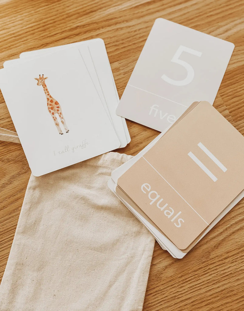 Warm wood background with Number Flash Cards by Little Roglets in 3 piles, with canvas bag. One card is white with a giraffe in the middle and it says "1 tall giraffe", other card is grey with a big white 5 in the middle, and the word "five" underneath, and last card is light brown with a "=" and the word "equals" in white.