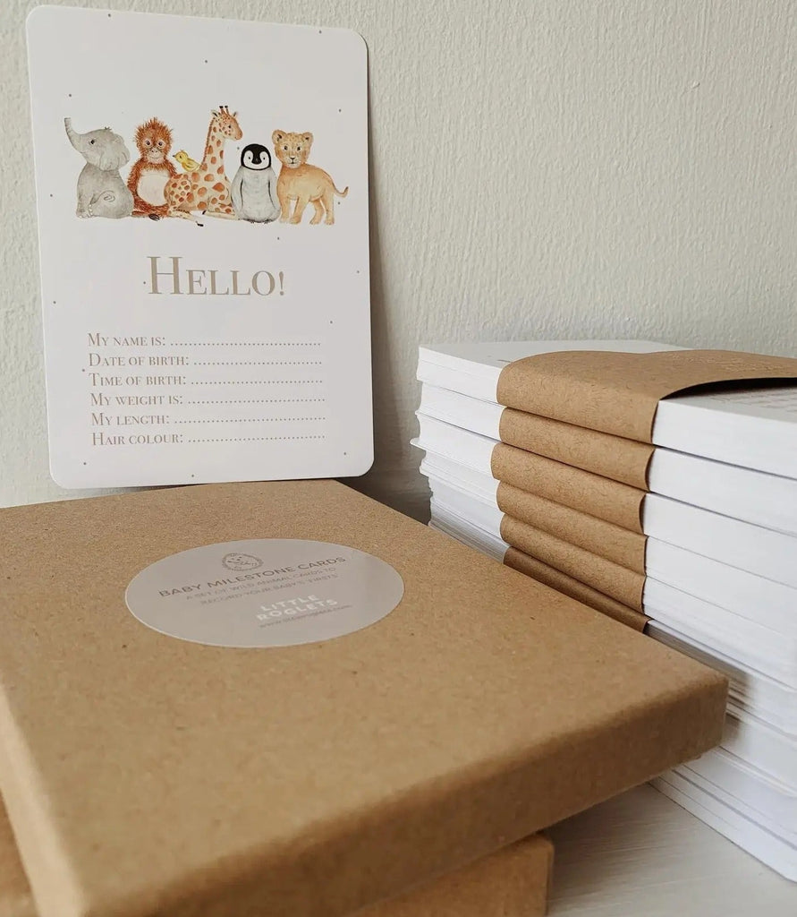White wall with 2 stacks of Milestone Card Set by Little Roglets, and 1 card on top. Card on top is white with baby animals in a line and says "Hello" with spots to put baby's birth stats.