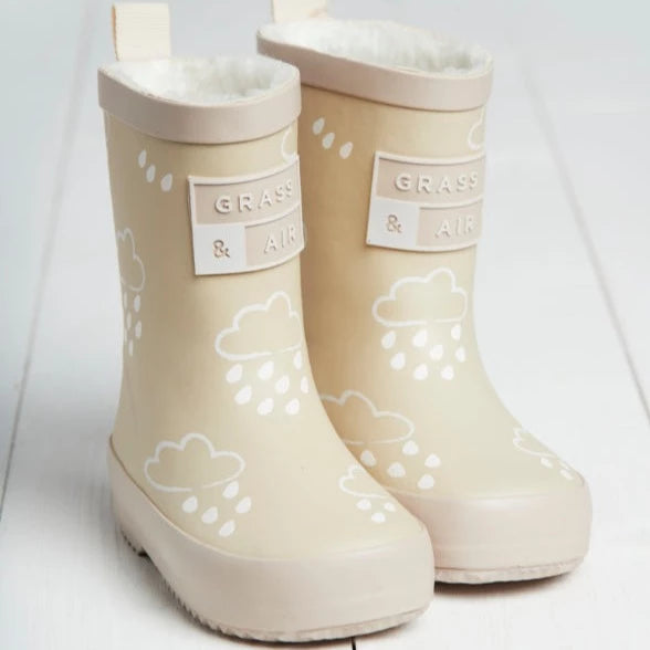 White background with Colour Changing Rain Boots in Stone by Grass & Air. Rain boots are a cream colour with white rain clouds all over, a fluffy interior, and a tag that says "Grass & Air".