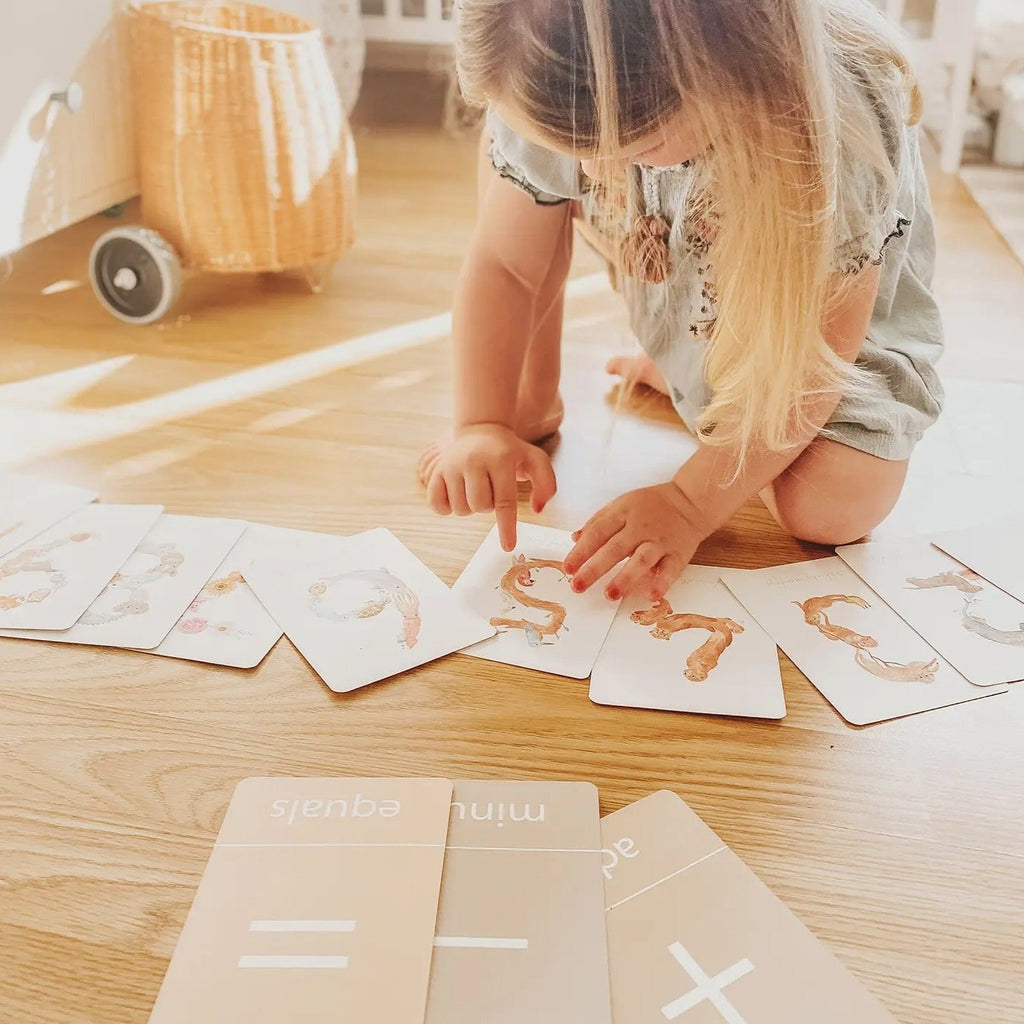 Wood floor with little girl leaning down, playing with Number Flash Cards by Little Roglets, which are all laid out in a row. Each card is white with a large number in the middle.