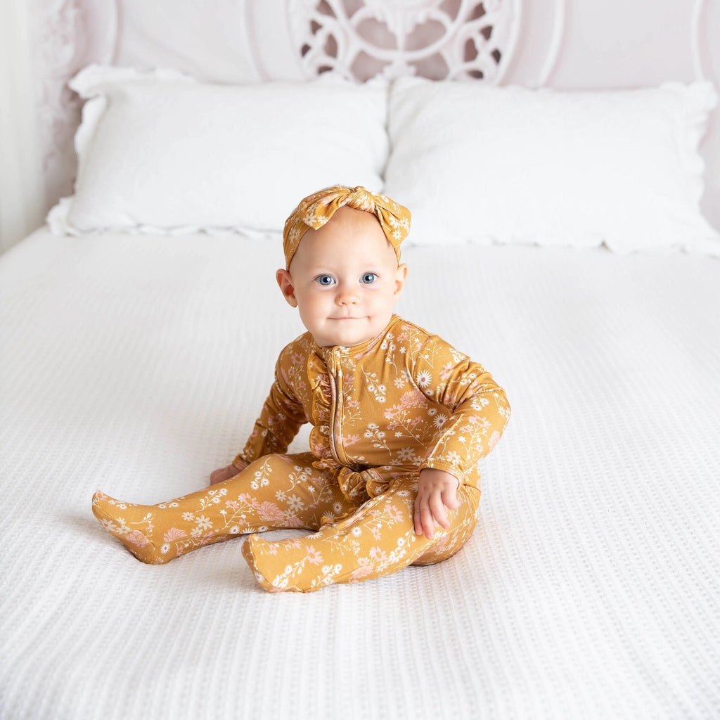 Straight on shot of baby girl sitting on bed wearing Bamboo Footie Sleeper in Mustard Floral by Emerson and Friends.