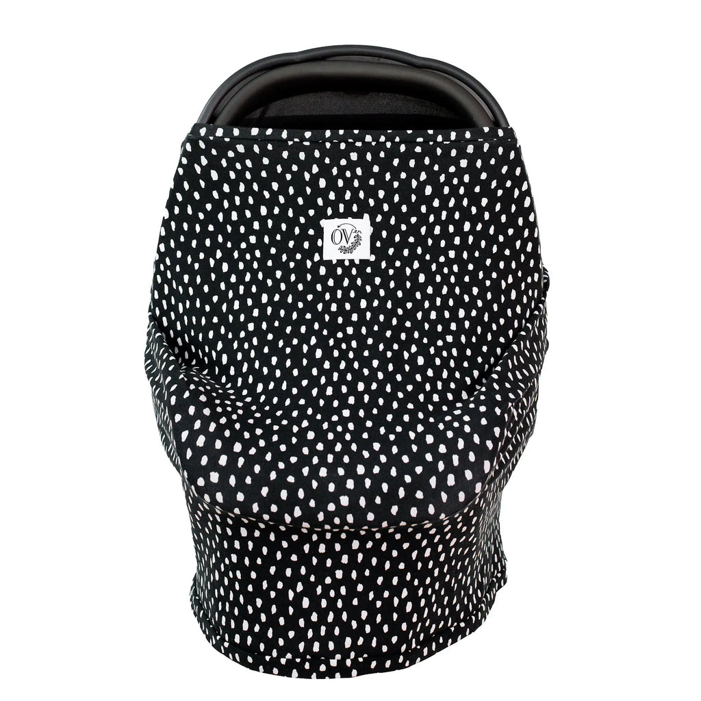 White background with a carseat and The Over Cover in Kelsey by The Over Company is on her car seat. This cover is black with white spots colour, with a white tag on the front with black font that says “OV” with greenery.