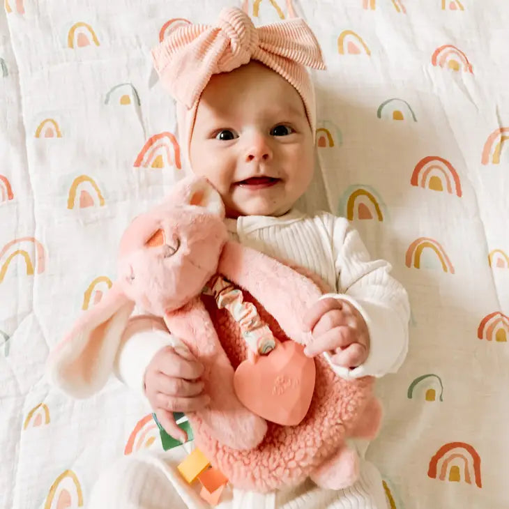 Baby model holding Itzy Lovey™ | Activity Plush & Teetherin Ana the Bunny by Itzy Ritzy. Model is wearing a cream sleeper and pink bow, laying on a swaddle with multi-coloured rainbows. 