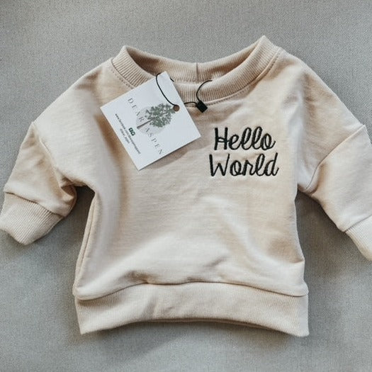 Grey background with Hello World Crewneck in Cloud by Dear Aspen. Crewneck is a cream colour with "Hello World" embroidered in black on the left chest.
