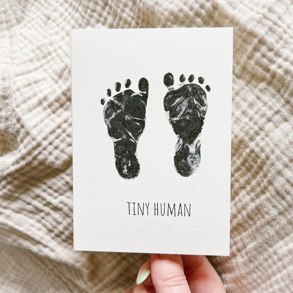 Tiny Human | Baby card by PARKES & bash being held by a hand in front of a grey/beige swaddle. 