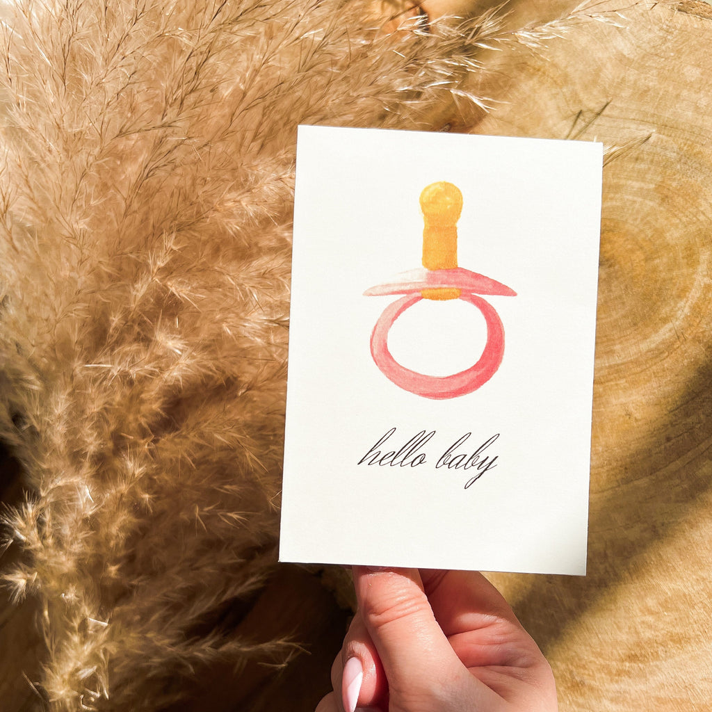 Hello Baby | Pink Paci card by Parkes & Bash, being held in front of wood and grass pampas with a hand holding it up. 