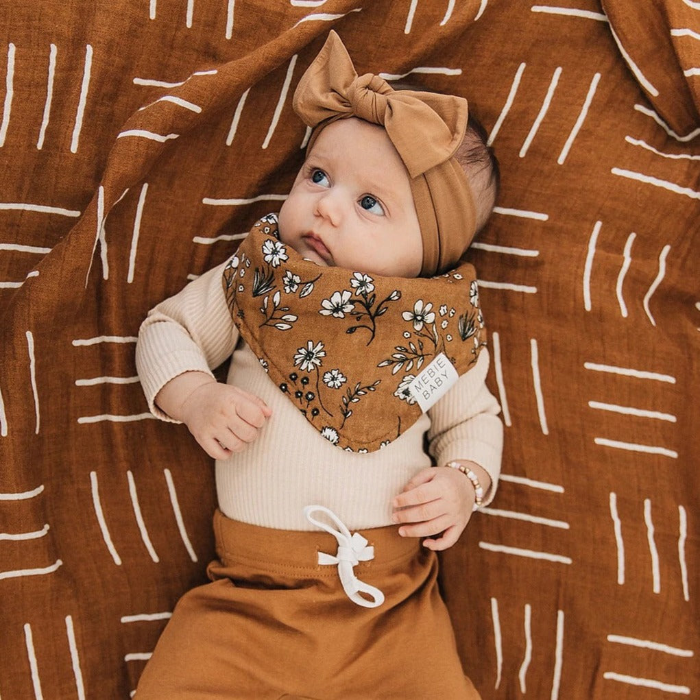 Overhead view of baby girl laying down, rust mudcloth swaddle underneath her, and she's wearing a Vintage Floral Bib by Mebie Baby. Triangle Bib is rust with white and black flowers.