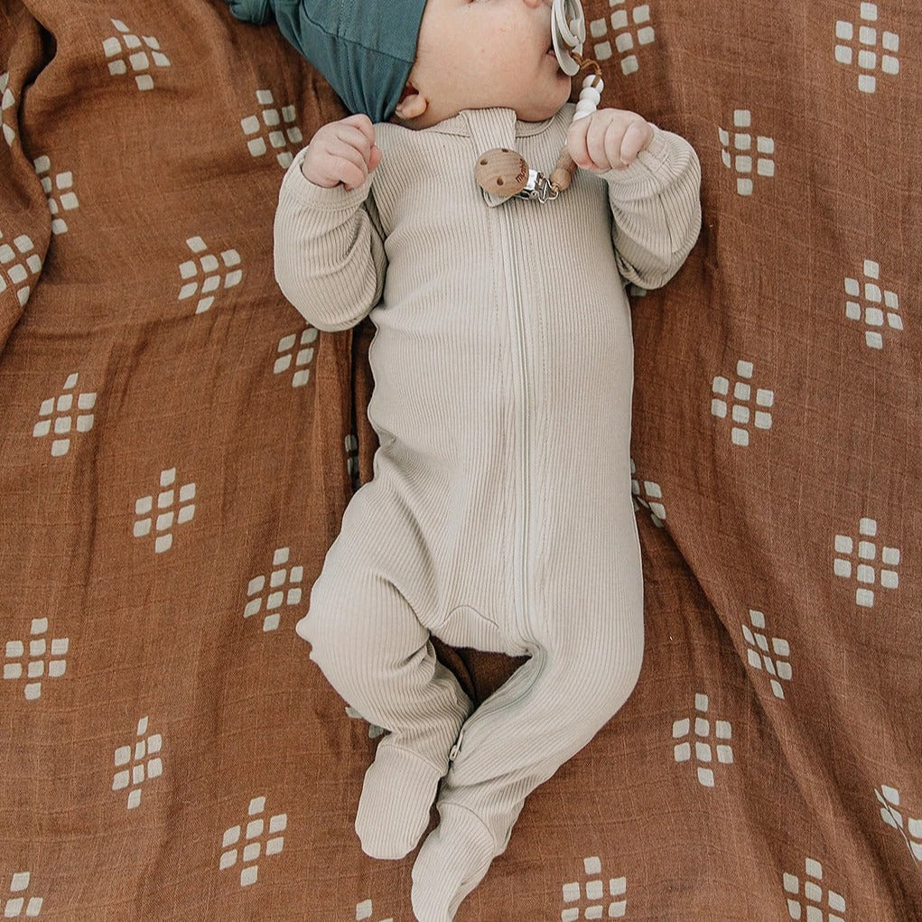 Overhead view of a baby laying on a Chestnut Textile Swaddle, wearing the Oatmeal Organic Ribbed Footed Zipper One-Piece by Mebie Baby.