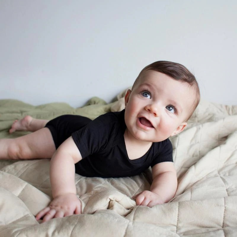 White background with bed, and blanket, and baby laying on stomach in Bodysuit in Midnight by Kyte Baby. Bodysuit is black.
