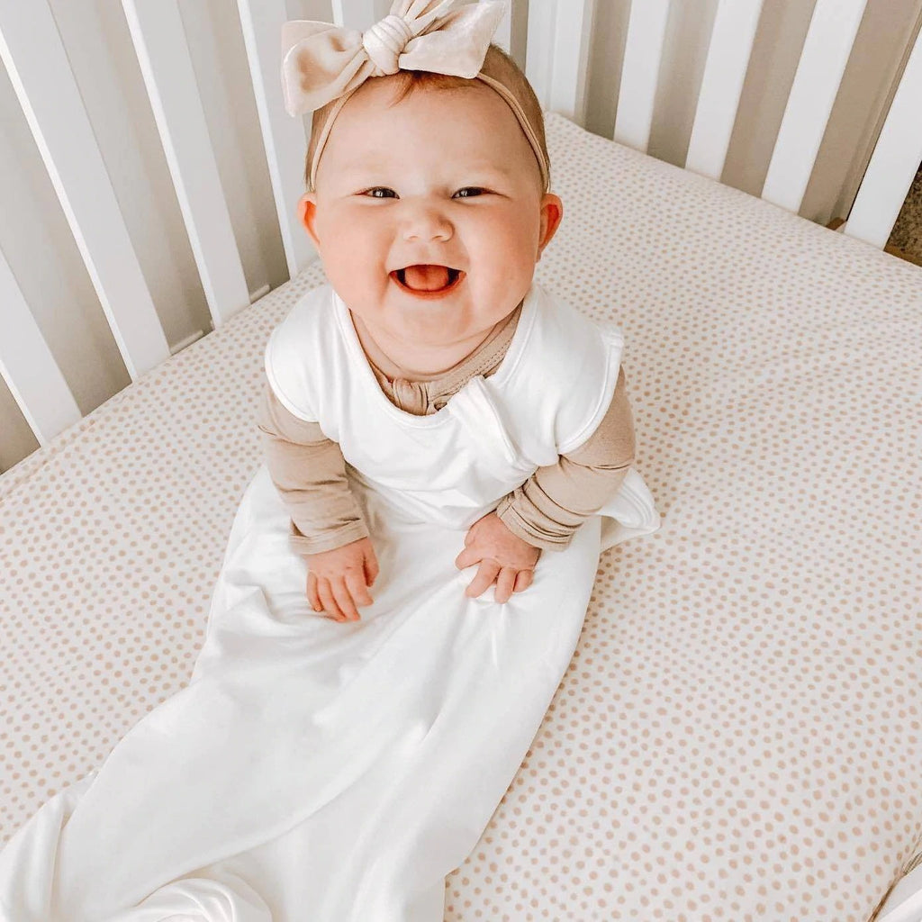 Overhead shot of baby in bed, sitting up and laughing, wearing a Sleep Bag .5 Tog in Cloud by Kyte Baby. Sleep bag is white, with a side zipper.