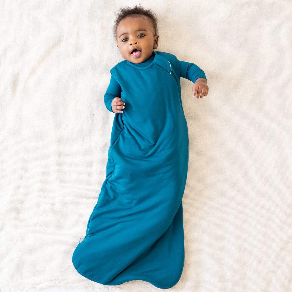 White background with baby laying down, tongue out, wearing a Sleep bag 1.0 Tog in Baltic by Kyte Baby. Sleep bag is a vibrant deep sea blue, with a zipper all the way down the side.