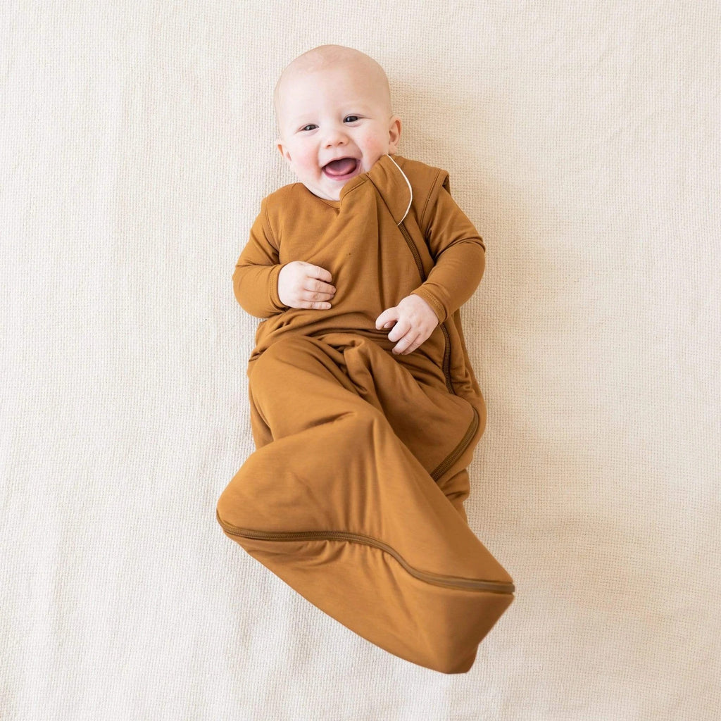 White background with overhead view of baby laying down, wearing a Sleep Bag 2.5 Tog in Nutmeg by Kyte Baby. Sleep bag is a light rust, with a side zipper going all around the bottom.