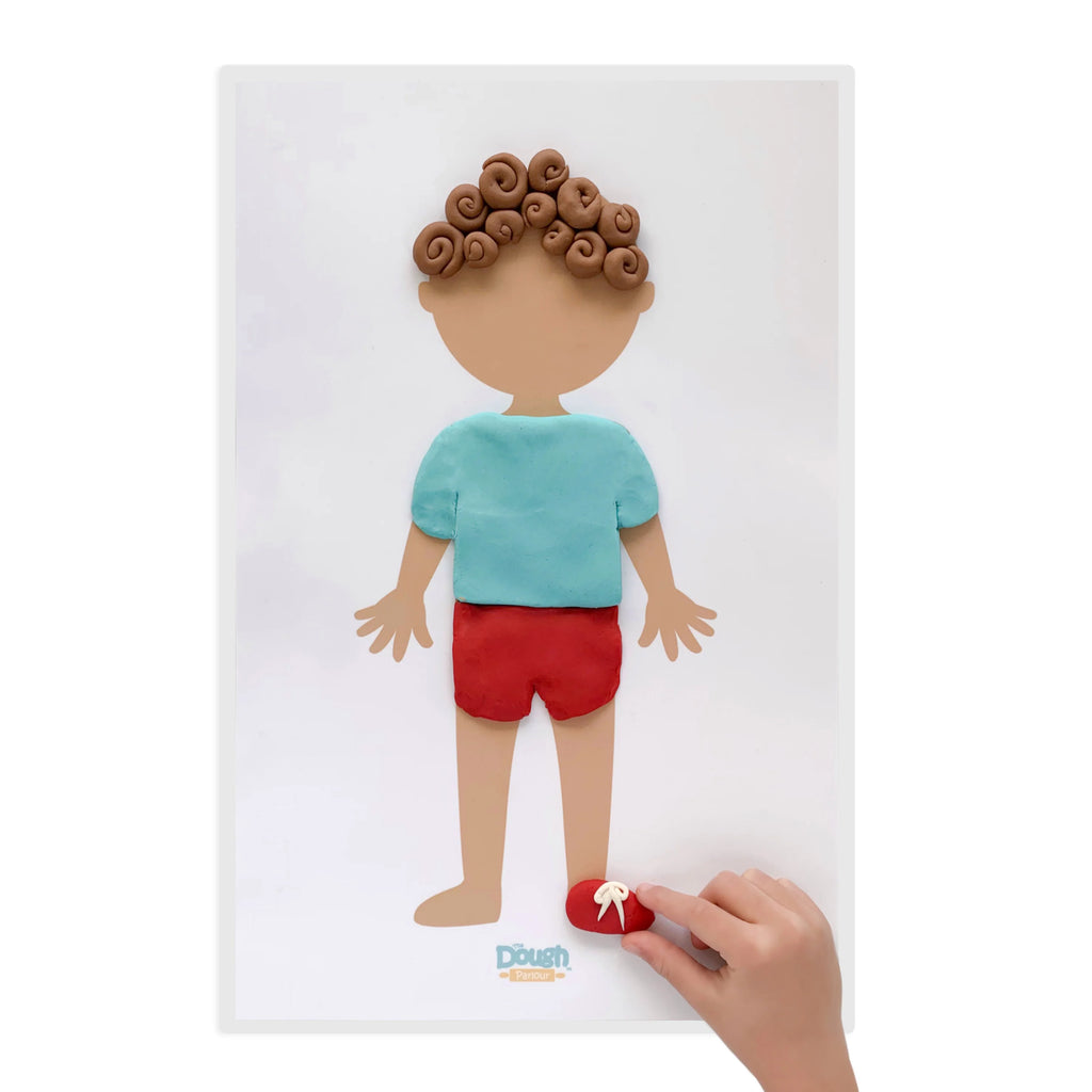 White background with a Creativity Playmat by Dough Parlour, and a hand molding play dough. Mat is of a person, and they have made dough to look like clothes, hair, and shoes.