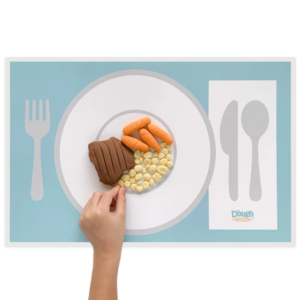 White background with a dinner setting playmat by Dough Parlour. Hand reaching out, making food out of play dough on the plate. 