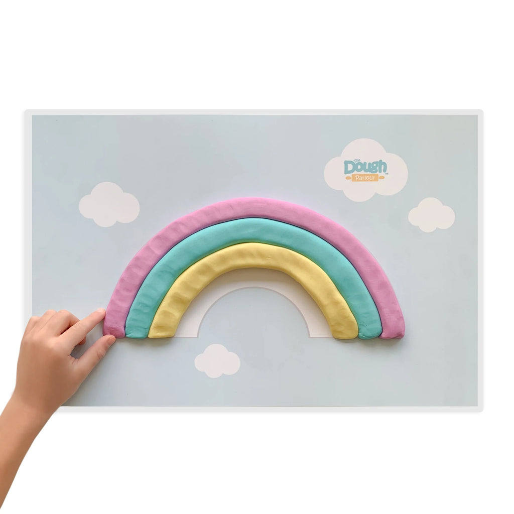 White background with Playmat by Dough Parlour. Playmat is of a rainbow, and a hand is molding play dough into a rainbow.