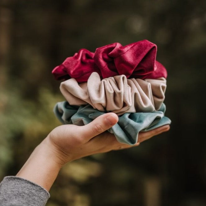 Hand holding a stack of Jumbo Velvet Scrunchies by Restored Signs & Decor, infront of some trees.