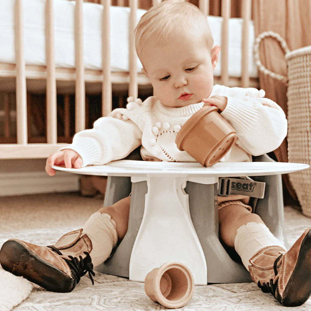 Close up of baby girl sitting in a Baby Chair & Booster Seat with tray by Upseat on the floor. Chair is grey with a white tray, and baby has stacking cups to play with.